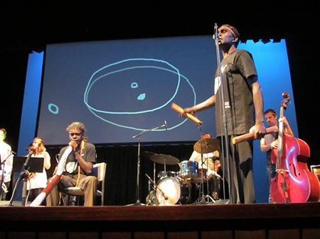 Daniel Wilfred, Benjamin’s brother, leads David Wilfred (didjeridu), Niko Schäuble (drums) and Philip Rex (double bass) as he sings the songs of Lutunba, a sacred site in Blue Mud Bay shown by the miny’tji projected on screen (illustrated by Ren Walters)